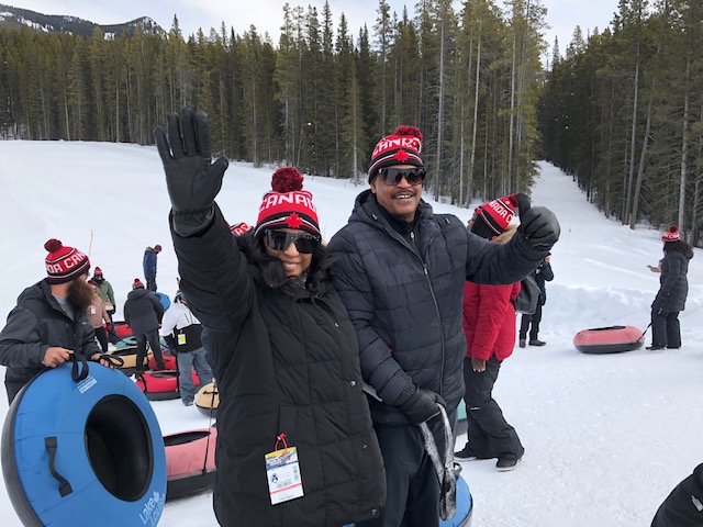A man and woman wave at the camera while standing in front of snow sleds.