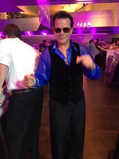 A man in blue shirt and black vest standing on dance floor.